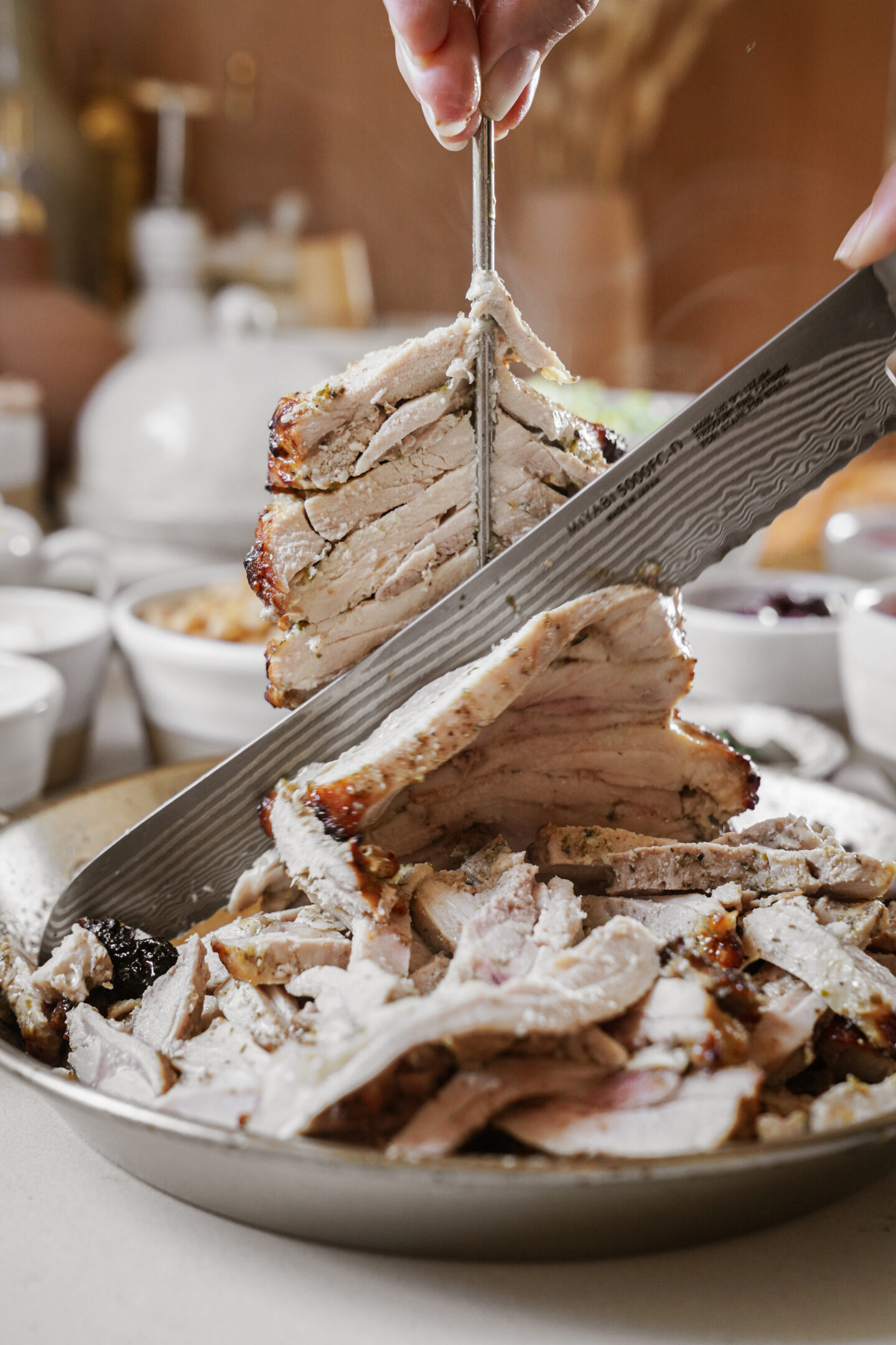 A hand uses a large, intricately patterned knife to slice through a stack of seasoned, roasted meat. The meat is served on a shallow beige plate, with small bowls of condiments in the blurred background.