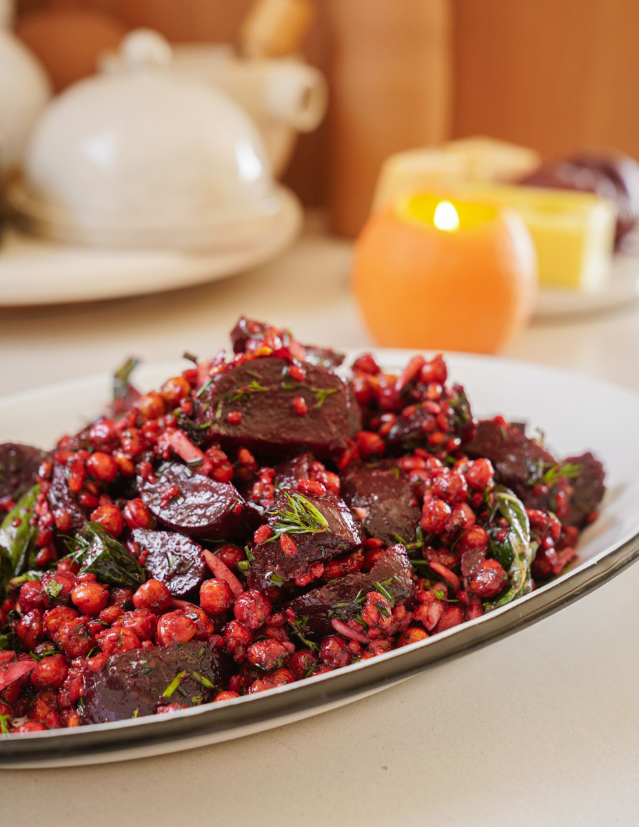 A white plate with a colorful salad featuring beets, grains, and leafy greens. The setting includes a blurred background with a lit candle and kitchen items, enhancing the cozy atmosphere.