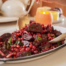 A platter of beet and pomegranate salad with fresh greens. Spoons are placed in the salad, suggesting serving. In the background, a lit candle and a butter dish are visible on the table.