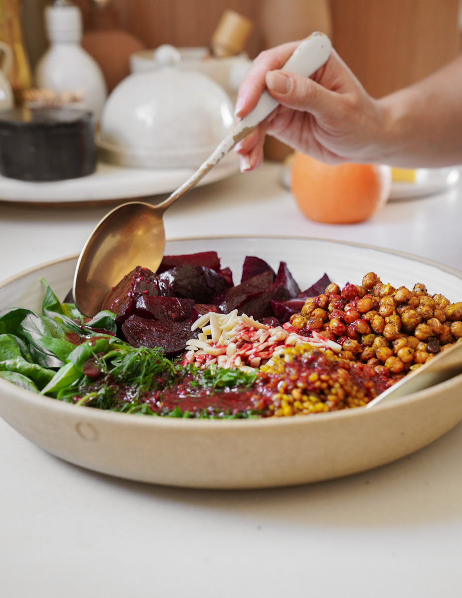 A hand holds a spoon over a bowl filled with vibrant ingredients: beetroot, roasted chickpeas, leafy greens, seeds, and grains. The background shows kitchen items and an orange.