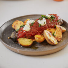 Grilled fish topped with a red sauce and dollops of cream, garnished with herbs, served on a plate with roasted potato halves.