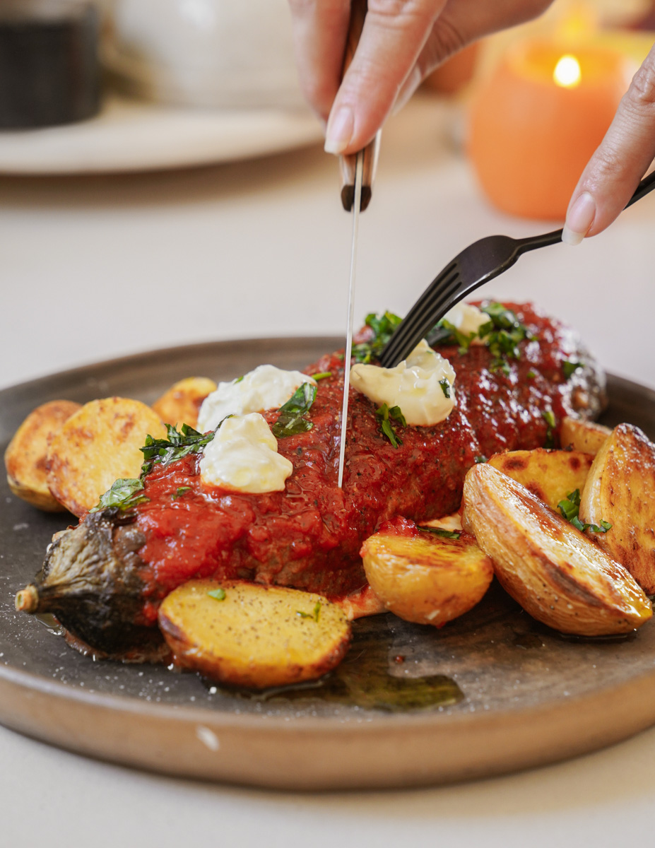 A person cutting into a roasted stuffed eggplant topped with tomato sauce and yogurt, garnished with herbs. Sliced roasted potatoes accompany the dish on a dark plate.