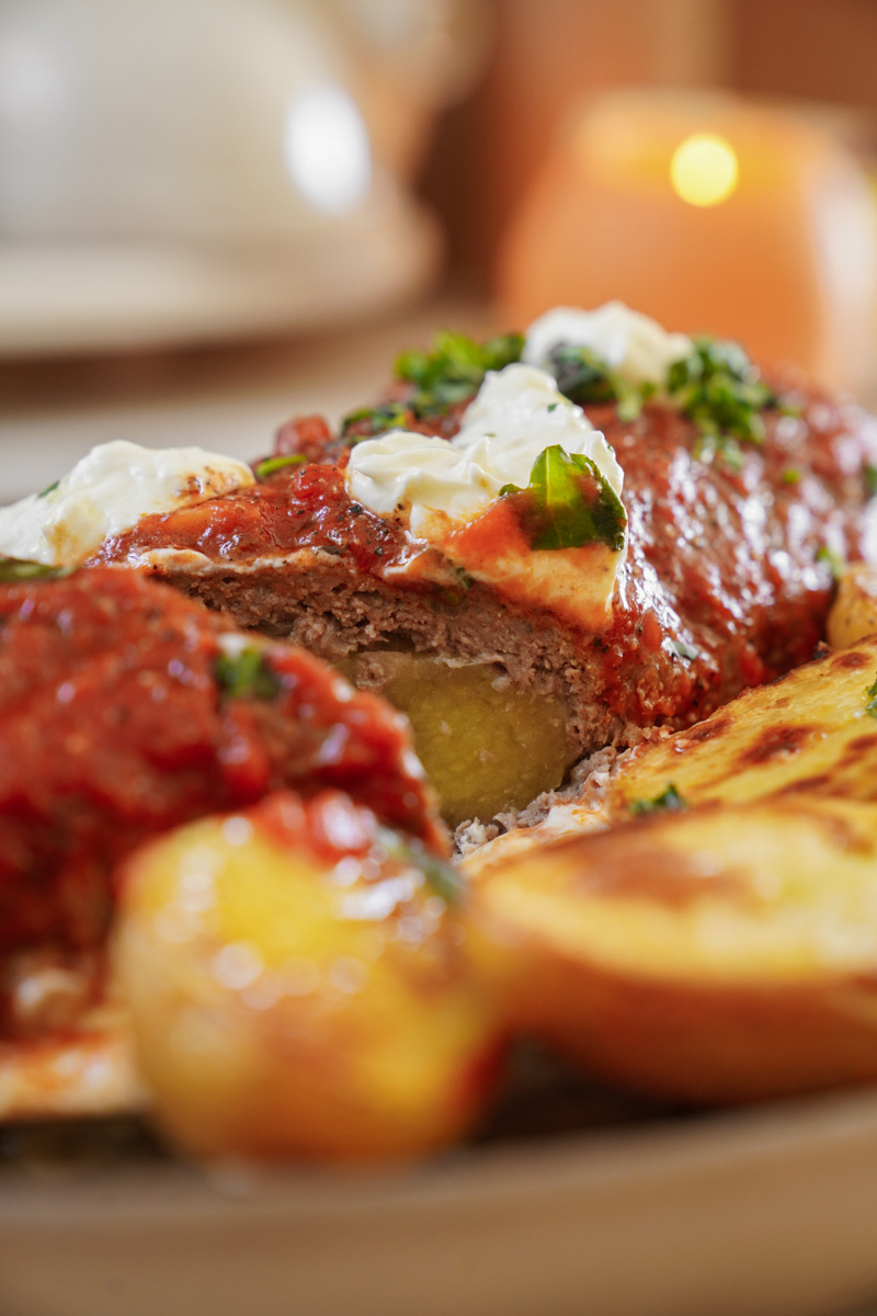 Close-up of a stuffed meatloaf topped with red sauce and dollops of creamy white topping, garnished with herbs. Its served with roasted potatoes on a plate. Warm, ambient lighting enhances the appetizing presentation.