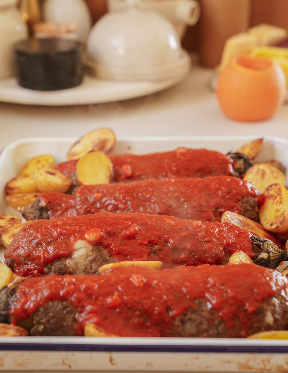 A baking dish filled with roasted vegetables and meatloaves covered in a rich tomato sauce. The dish is surrounded by kitchen items, including a teapot, a spice container, and a peeled citrus fruit, all set on a light countertop.