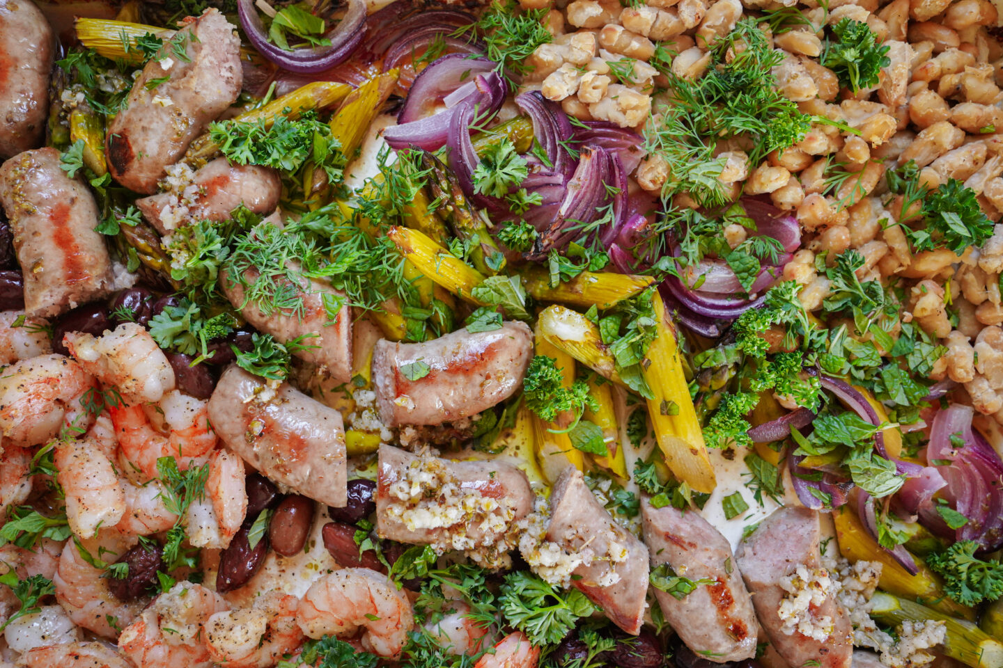 A colorful assortment of sautéed shrimp, sausages, red onions, chickpeas, olives, and asparagus, all garnished with fresh herbs like parsley. The vibrant mix showcases a variety of textures and flavors.