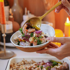 A hand holds a ceramic bowl filled with a colorful seafood and vegetable dish, garnished with herbs. A golden spoon lifts a portion of the meal. Soft candlelight glows in the background, creating a cozy atmosphere.