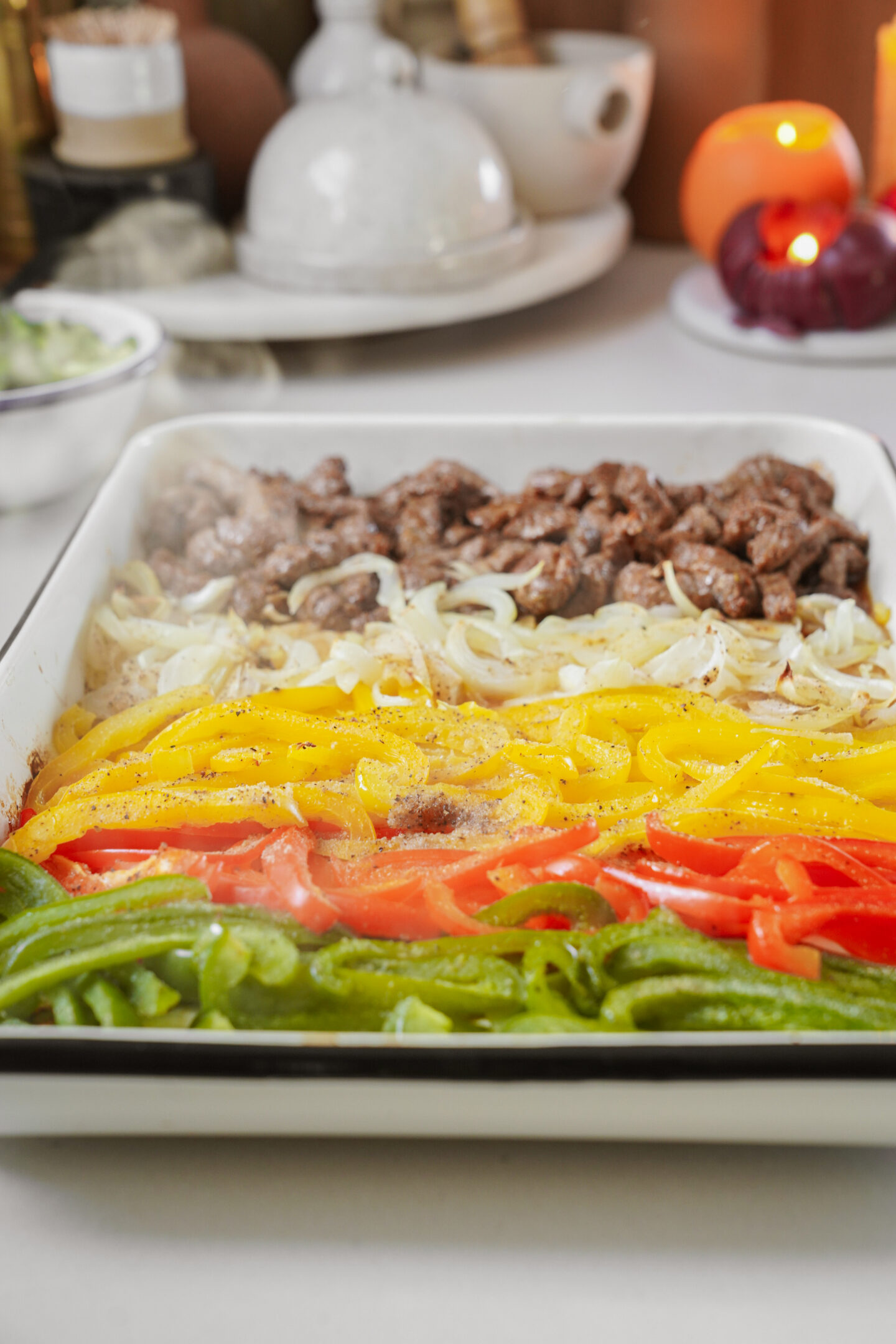 A colorful dish featuring sliced beef, onions, yellow peppers, red peppers, and green peppers arranged in rows on a rectangular tray. A steaming garnish is visible. Candles and crockery are in the blurred background.