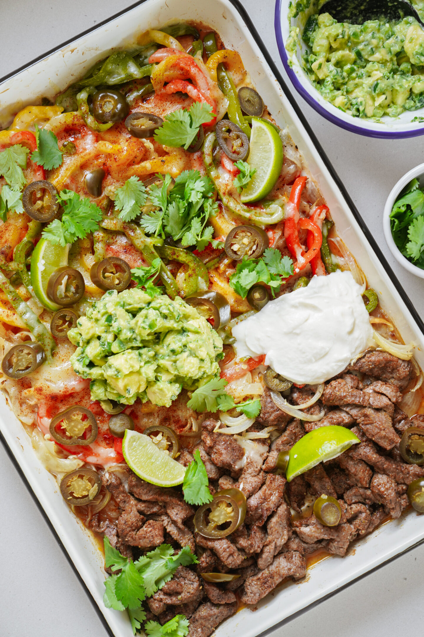 A colorful platter with sliced beef steak, bell peppers, jalapeños, and melted cheese. Garnished with lime wedges, cilantro, a dollop of sour cream, and a scoop of guacamole. A small bowl of guacamole is nearby.