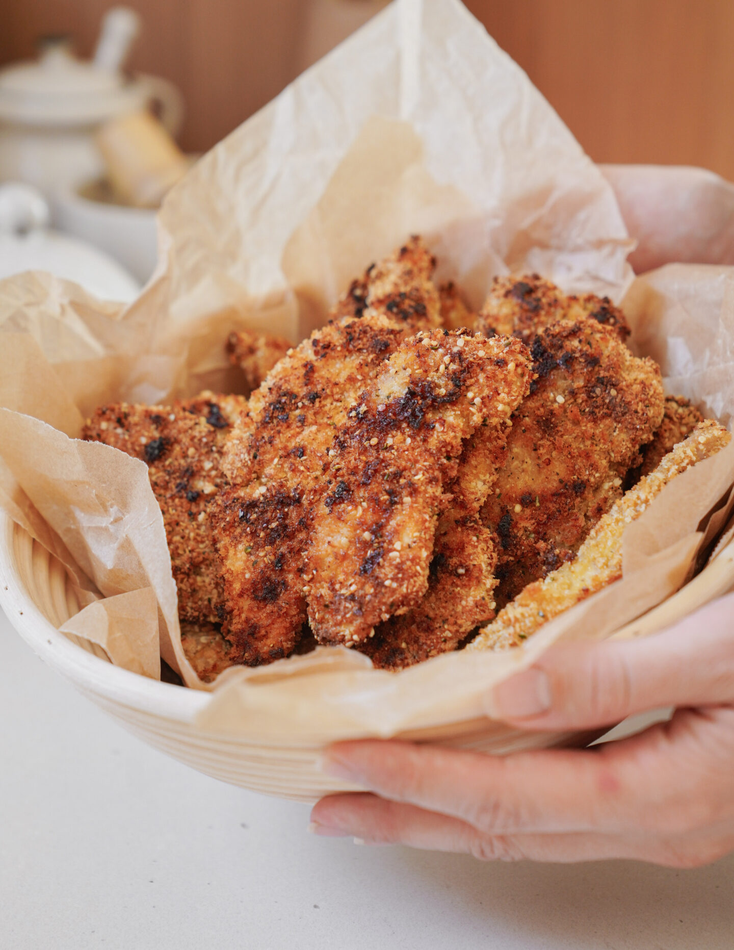 A hand holding a bowl lined with parchment paper, filled with crispy, golden-brown breaded chicken pieces. The chicken looks freshly cooked and is arranged appetizingly in the bowl.