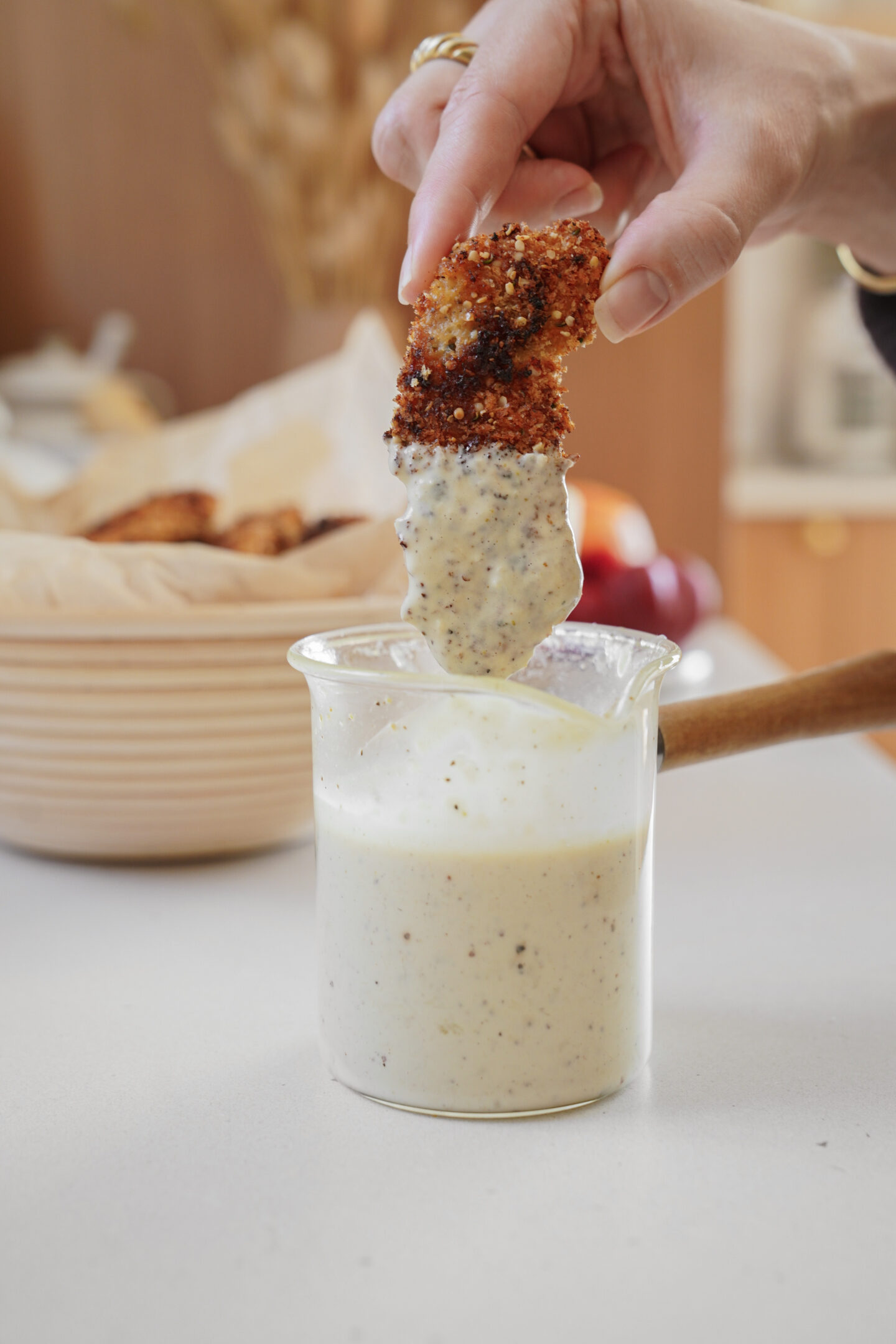 A hand is dipping a piece of breaded fried food into a clear jar filled with creamy white sauce. A basket of similar food pieces is partially visible in the background. The scene is set on a light-colored countertop.