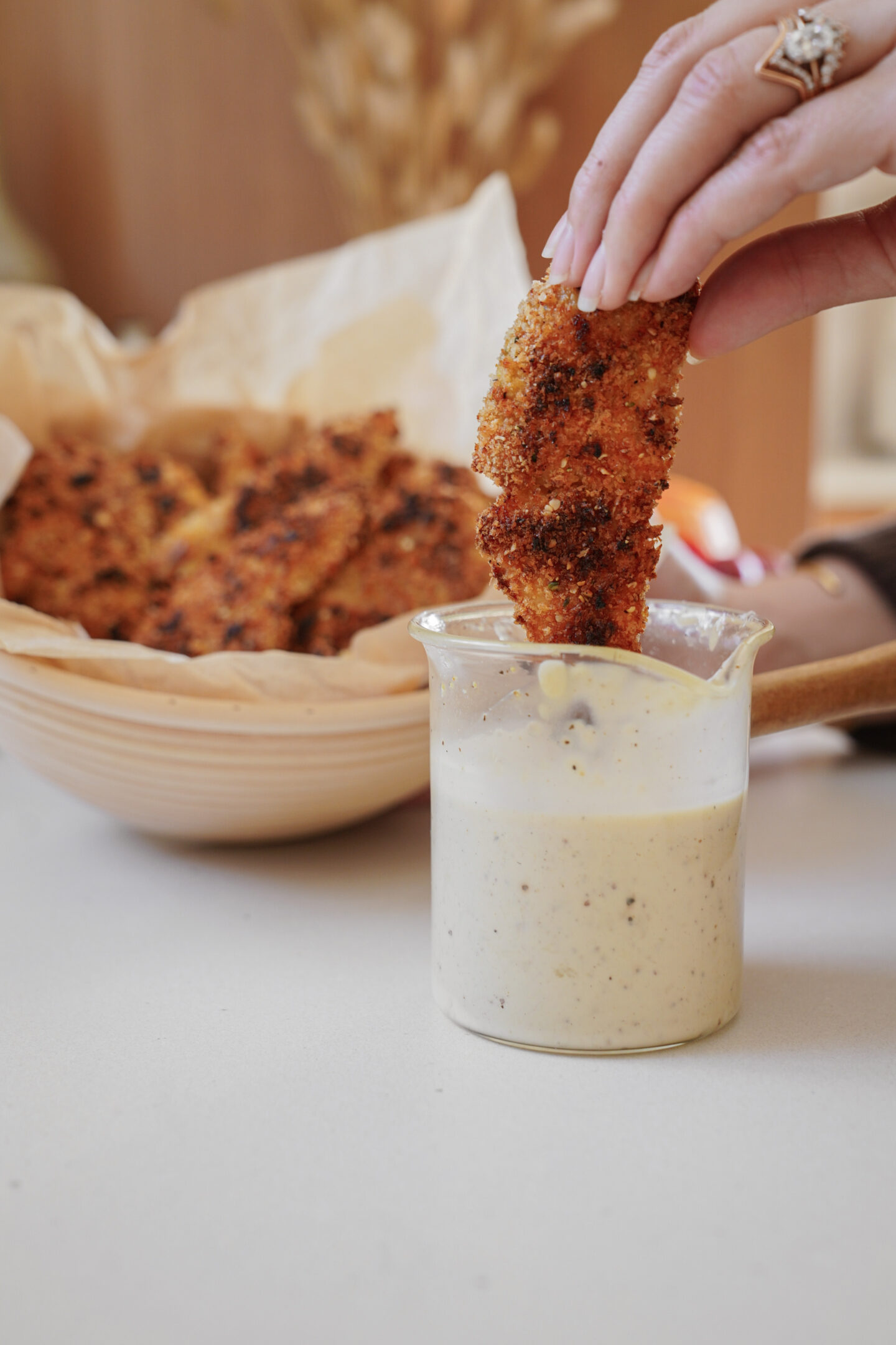 A hand with manicured nails dips a crispy, breaded chicken tender into a small glass of creamy sauce. In the background, more chicken tenders are stacked in a wooden bowl lined with parchment paper.