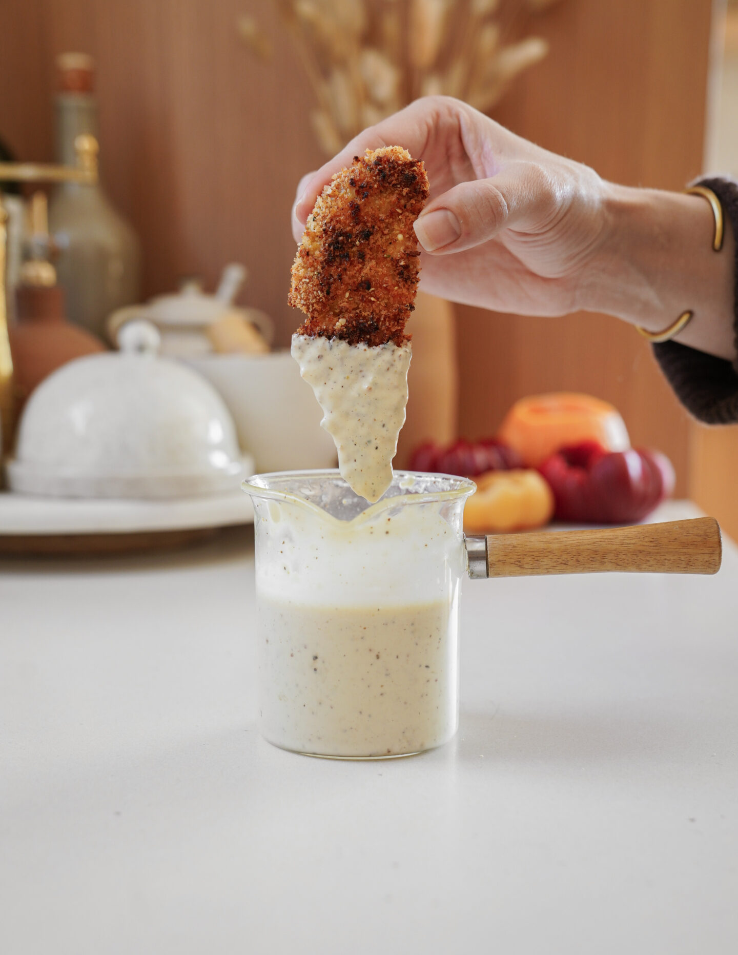 A hand dipping a breaded, fried snack into a small glass pot of creamy sauce with speckles. The pot has a wooden handle. In the blurred background, there are kitchen items like a butter dish and fruits.