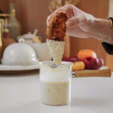 A hand is dipping a piece of breaded and fried food into a small glass pot filled with a creamy sauce. The pot has a wooden handle and is on a kitchen counter with blurred kitchen items in the background.