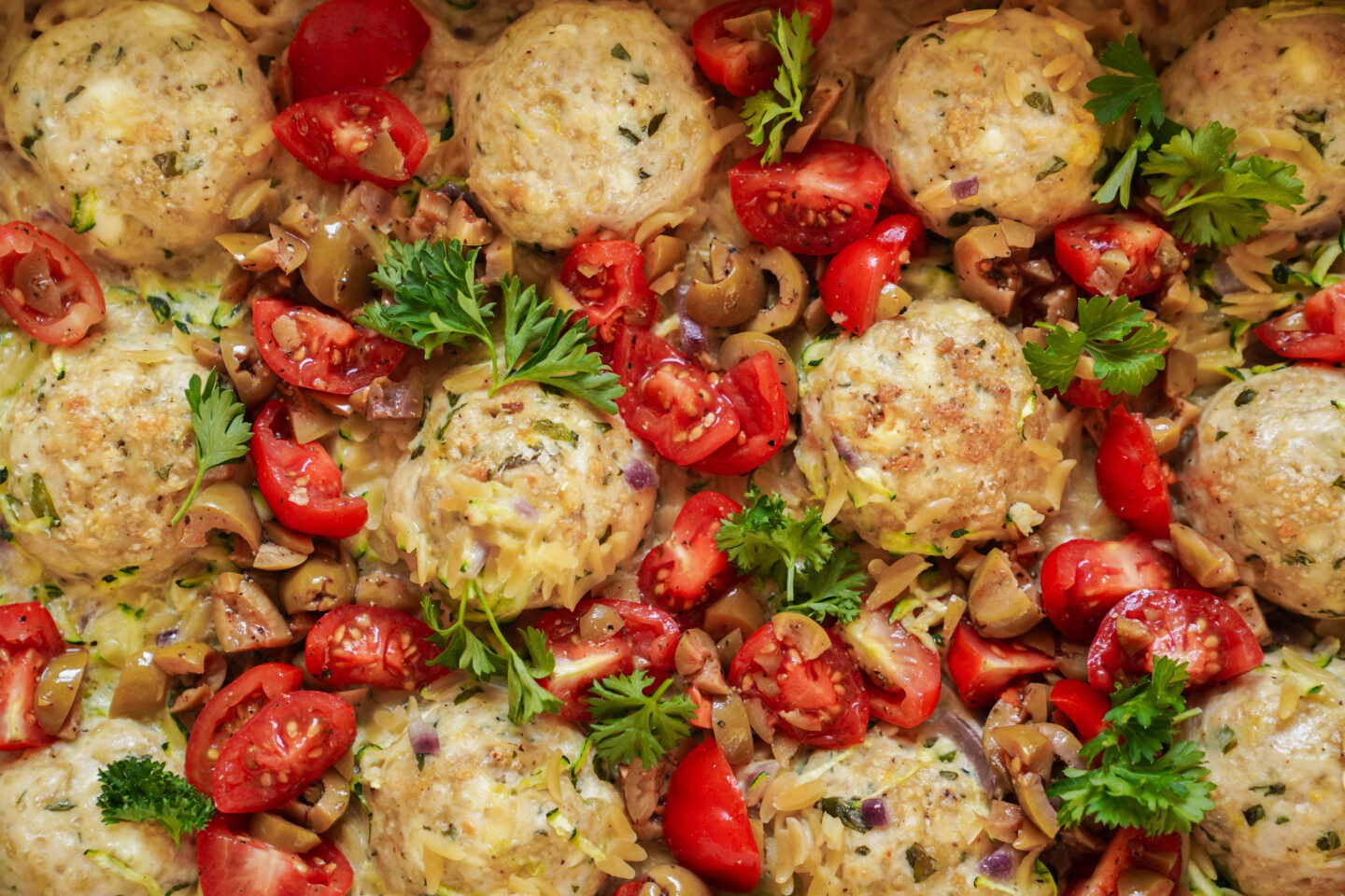 A close-up of a dish featuring meatballs garnished with chopped tomatoes, olives, and fresh parsley. The meatballs are cooked and surrounded by vibrant red tomatoes and green herbs, creating a colorful and appetizing presentation.