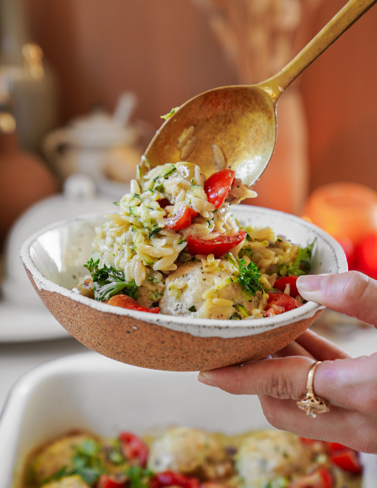 A hand holding a bowl of creamy orzo pasta mixed with shredded zucchini, cherry tomatoes, and garnished with fresh parsley. A gold spoon is seen serving the pasta. Warm lighting creates a cozy atmosphere in the background.