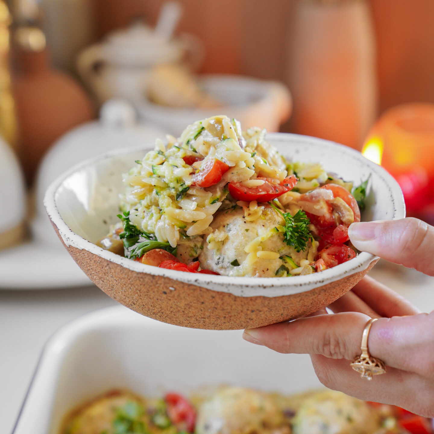 A hand holding a bowl of creamy orzo pasta with herbs, cherry tomatoes, and chunks of chicken. The dish is garnished with fresh parsley. Background shows a blurred kitchen setting with warm lighting.