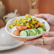 A person holds a bowl with white rice topped with cooked salmon chunks, sliced cucumbers, and a mango-avocado salsa. The background features soft, neutral tones with blurred elements like a teapot and decorative items.