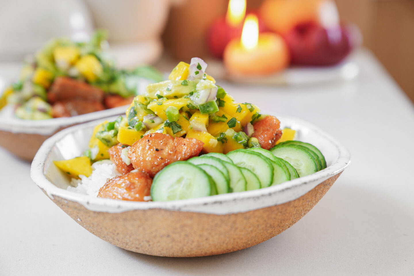 A bowl contains white rice, topped with slices of cucumber, grilled chicken pieces, and a colorful mango salsa made with cilantro and onions. Theres a blurred view of lit candles in the background.