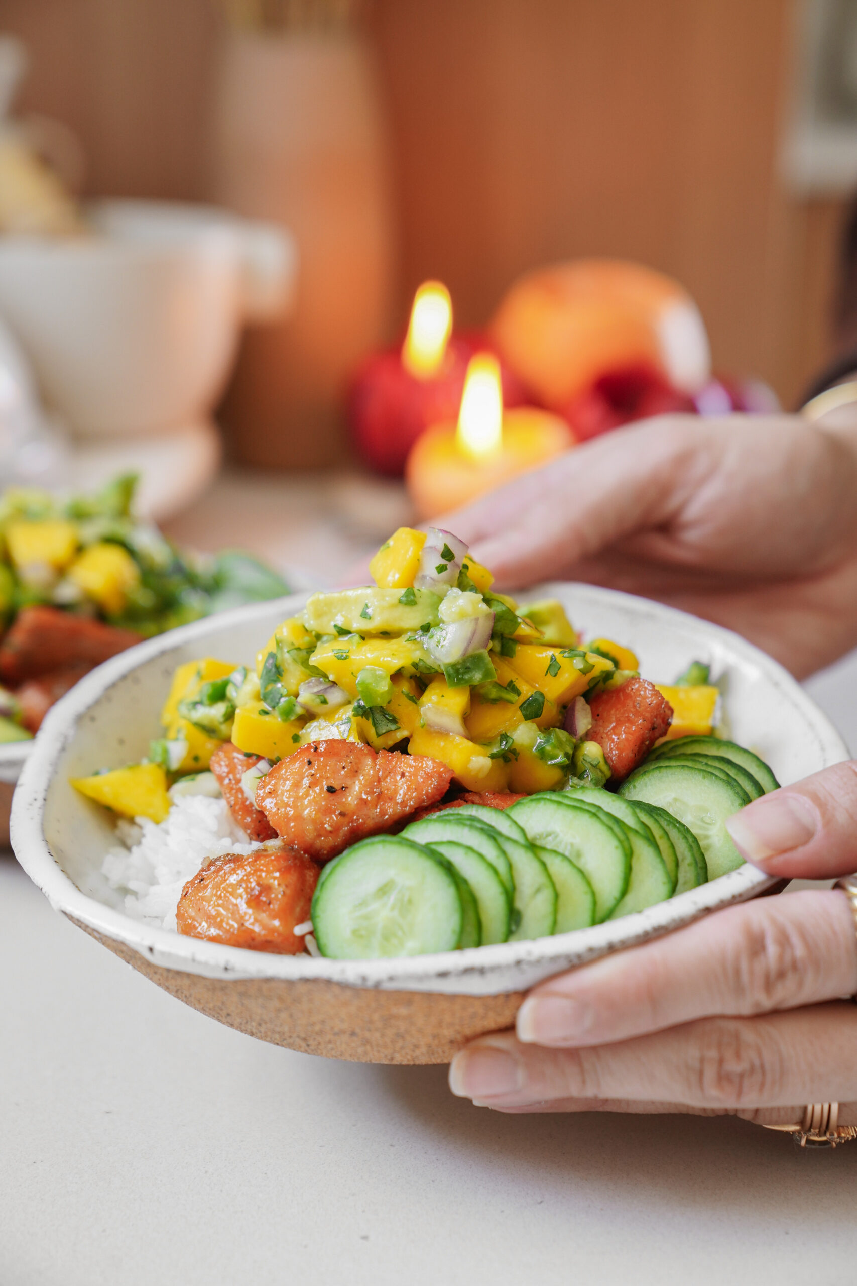 A person holds a bowl of food featuring sliced cucumbers, mango salsa, and glazed tofu pieces on a bed of rice. Lit candles and a warm, blurred background create a cozy atmosphere.