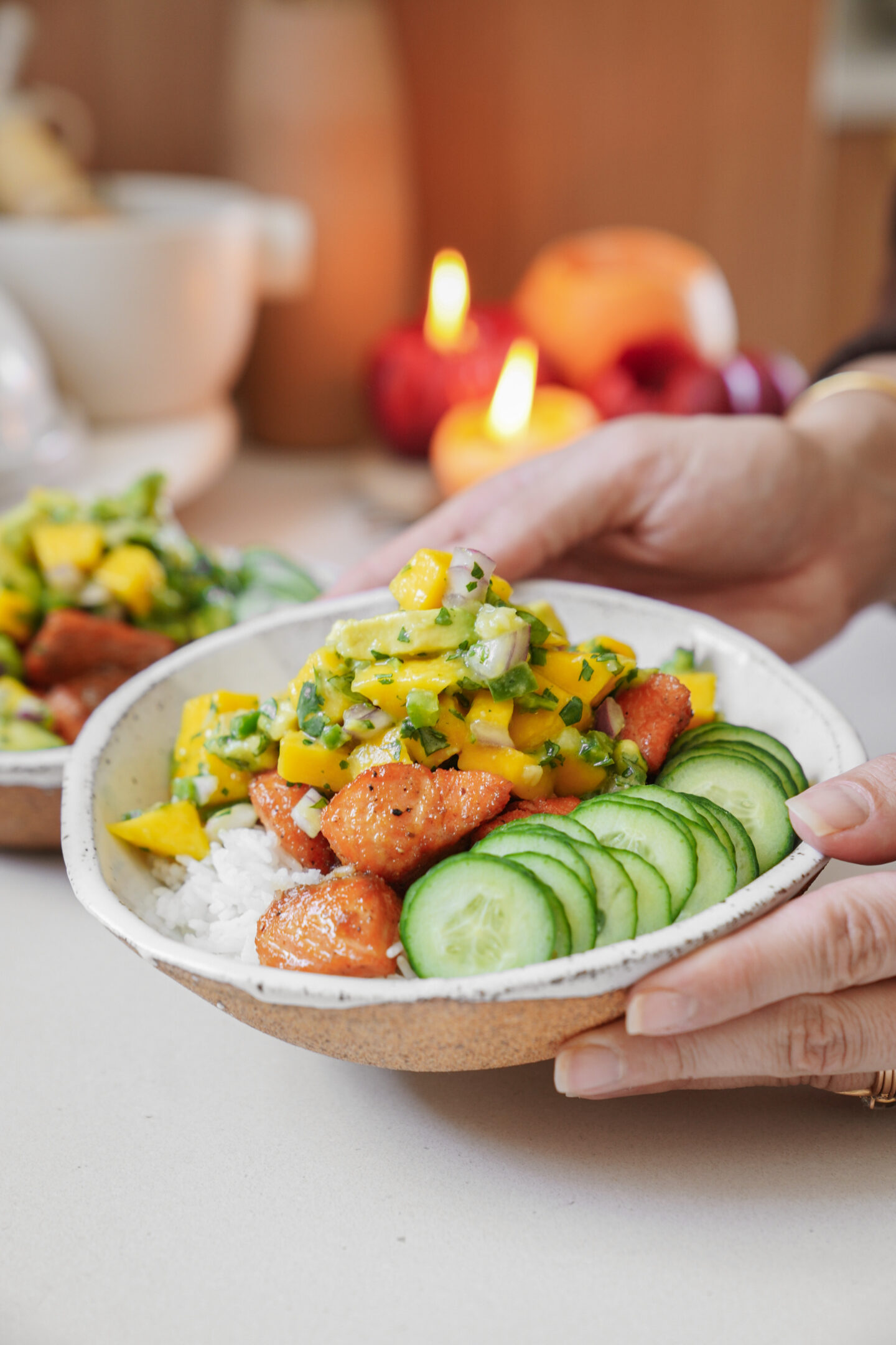 A person holds a bowl with sliced cucumber, diced salmon, white rice, and a mango salsa with herbs. Another similar bowl is in the background, along with lit candles, creating a cozy dining atmosphere.