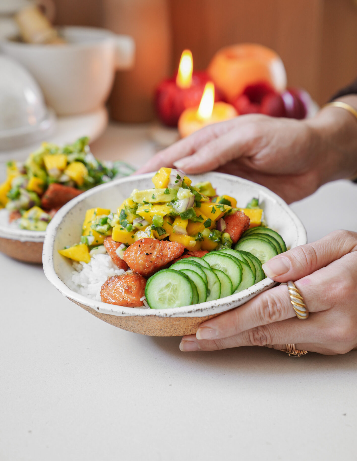 Hands holding a bowl of rice topped with cooked salmon, sliced cucumbers, and mango salsa. Another similar bowl is in the background. Candles and blurred fruit are visible in the warm, softly lit setting.