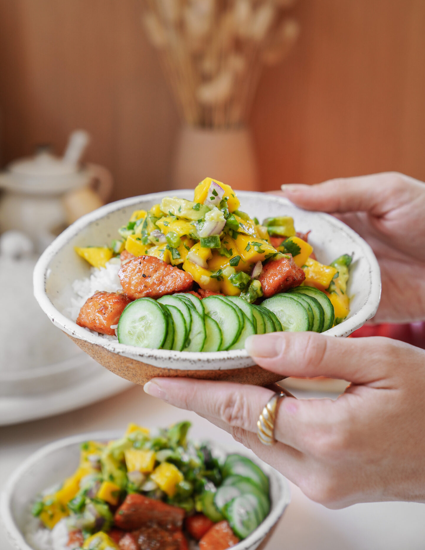 A person holds a bowl of rice topped with sliced cucumbers, diced salmon, and yellow mango salsa. Another similar bowl is in the background, and a teapot is on the table beside dried wheat stalks.