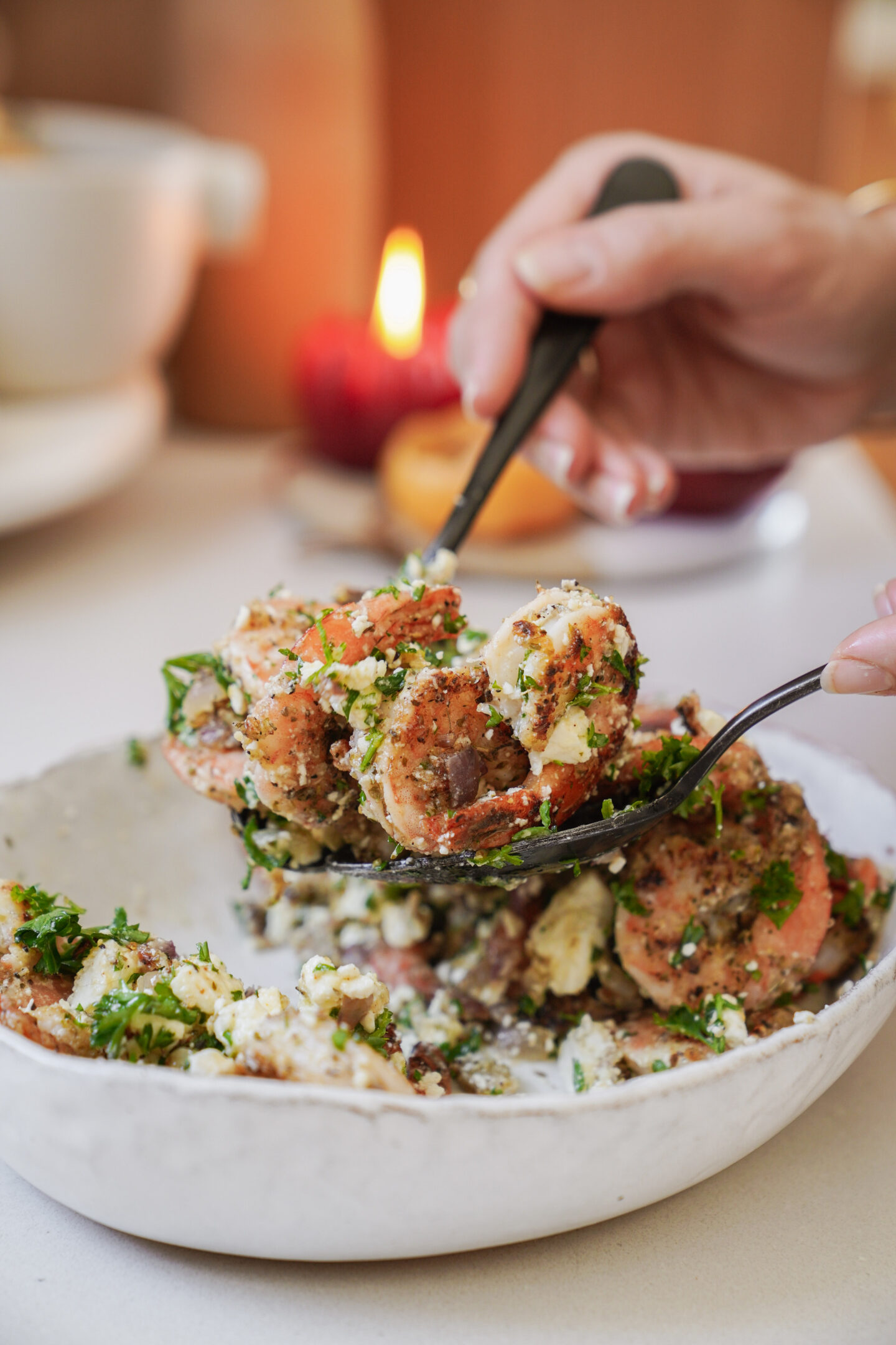 Close-up of a person using a fork and spoon to serve a dish filled with seasoned shrimp, herbs, and cheese in a white bowl. A candle is softly lit in the background, creating a warm ambiance.