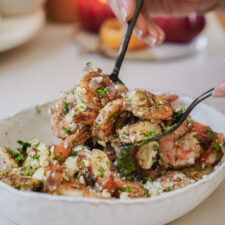 A person uses a fork to scoop a serving of grilled shrimp salad from a white bowl. The salad is mixed with crumbled cheese, chopped herbs, and diced vegetables. A blurred background shows more food and a lit candle.