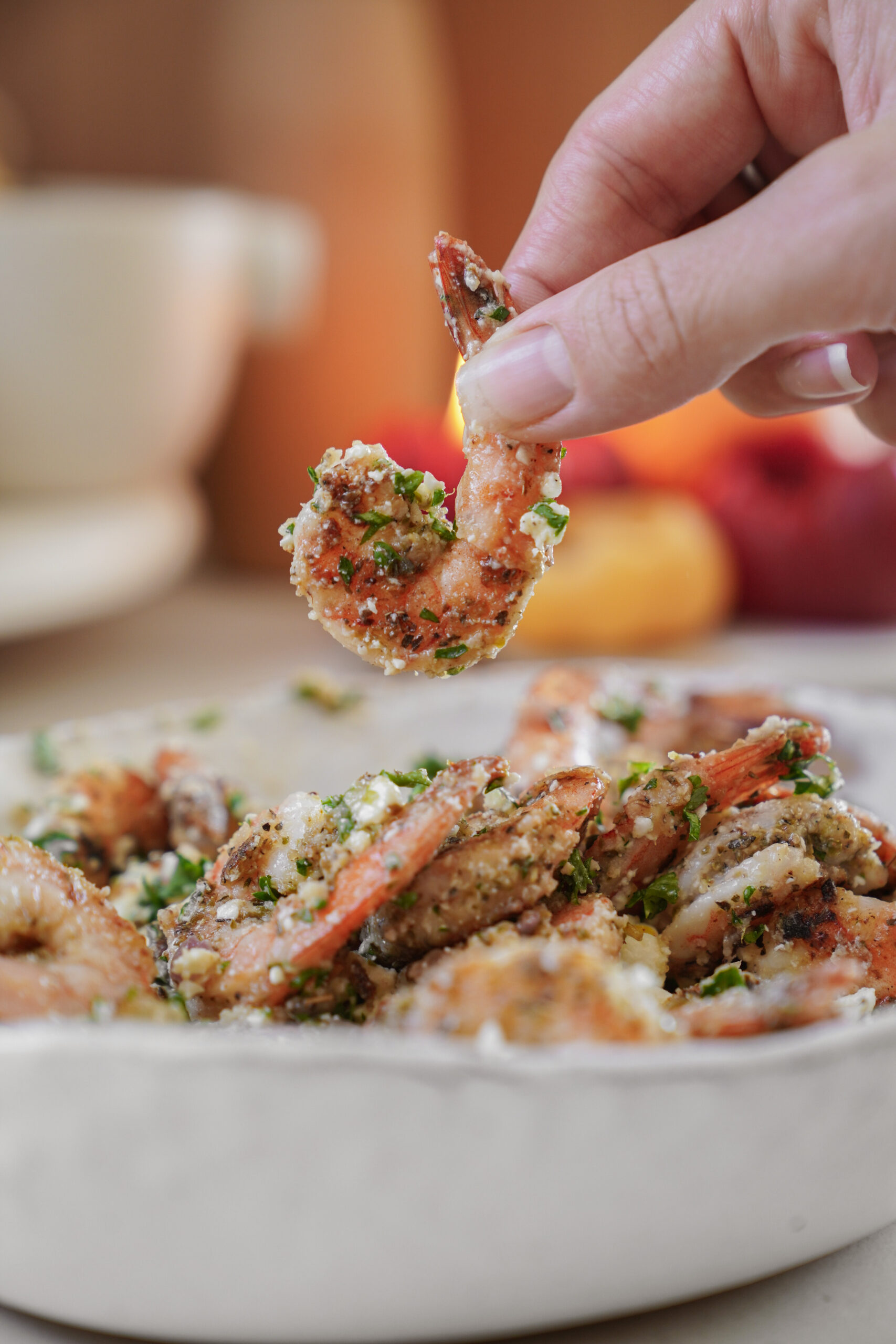 Close-up of a hand holding a seasoned, cooked shrimp above a bowl filled with more shrimp. The shrimp are garnished with herbs and spices, and the background is softly blurred, featuring a cup and fruit.