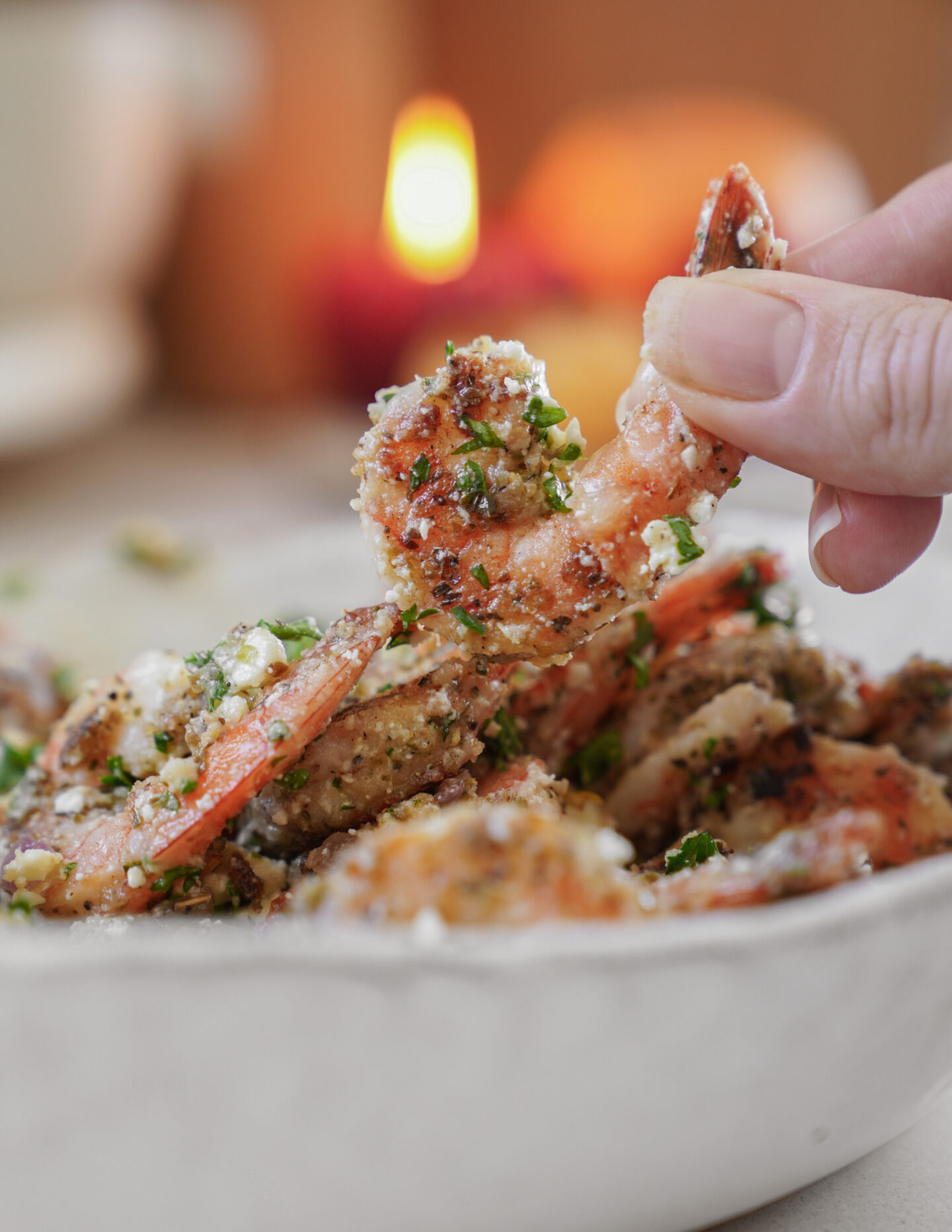 A close-up of a hand holding a seasoned shrimp over a bowl filled with more shrimp. The shrimp are covered in herbs and spices. A softly lit candle is blurred in the background, creating a warm ambiance.