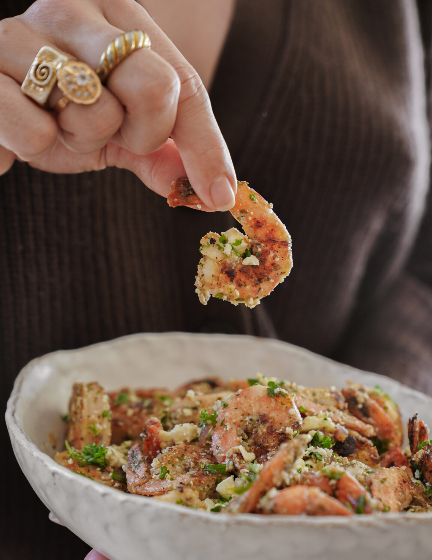 A person holding a seasoned shrimp above a bowl filled with shrimp and herbs, wearing a brown sweater and a gold ring with an intricate design. The shrimp is garnished with green herbs and crumbs.