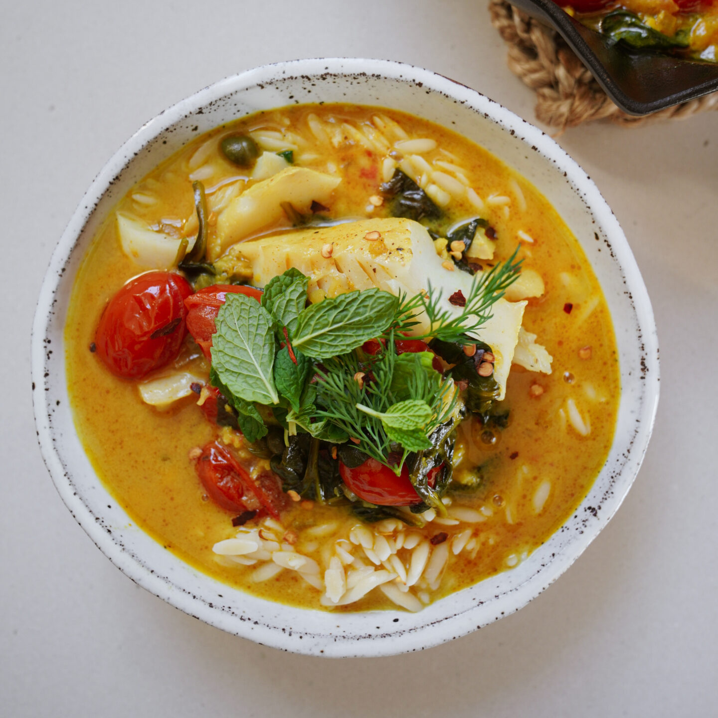 A bowl of vibrant yellow curry with fish, cherry tomatoes, spinach, and orzo pasta is garnished with fresh mint and dill. The creamy broth is topped with a sprinkle of red chili flakes, creating a colorful and appetizing presentation.