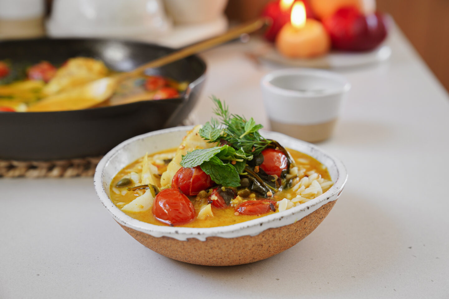 A ceramic bowl filled with yellow curry, cherry tomatoes, herbs, and rice is placed on a white surface. A skillet with more curry is in the background, beside candles and blurred objects.