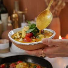 Hand holding a bowl of curry, garnished with fresh herbs and cherry tomatoes. A spoon is adding sauce to the dish. The background shows blurred kitchen items and a lit candle.