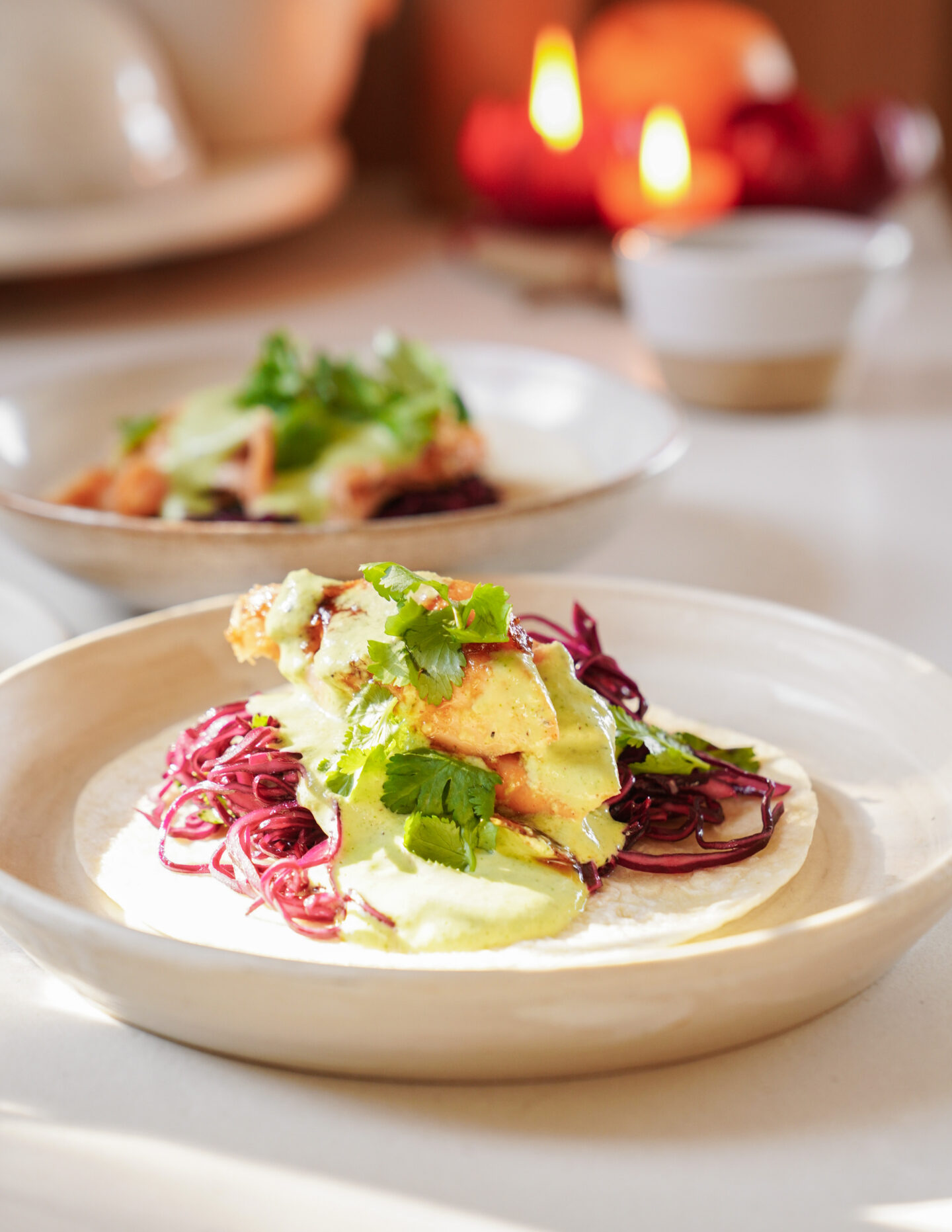 A plate of tacos on a white ceramic dish, topped with crispy fried chicken, shredded red cabbage, sliced cucumbers, and fresh cilantro. Two glowing candles and a small bowl are blurred in the background, creating a cozy ambiance.