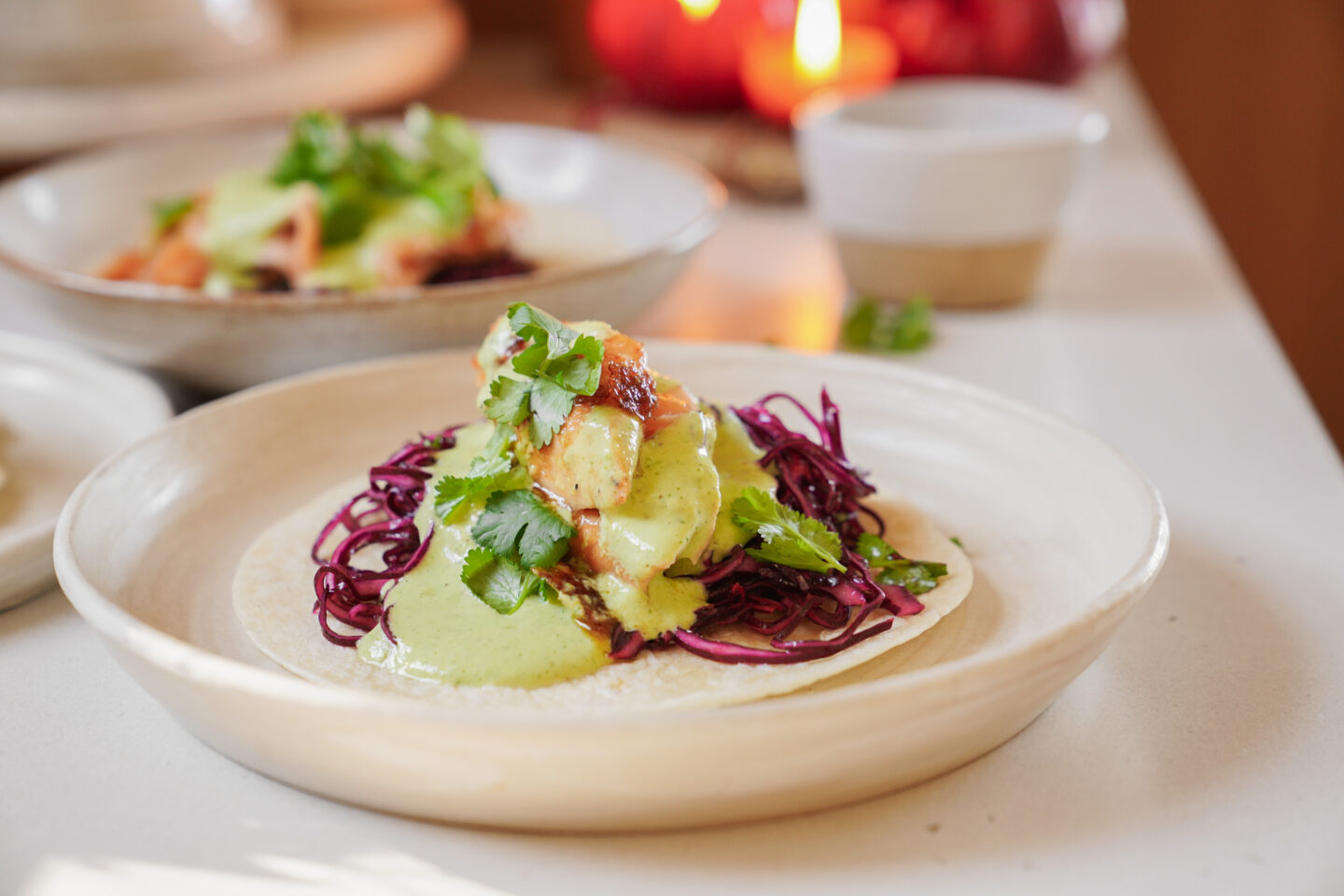 A taco on a white plate with grilled chicken, red cabbage, cilantro, and green sauce. The background features another plate, a candle, and a small white cup.