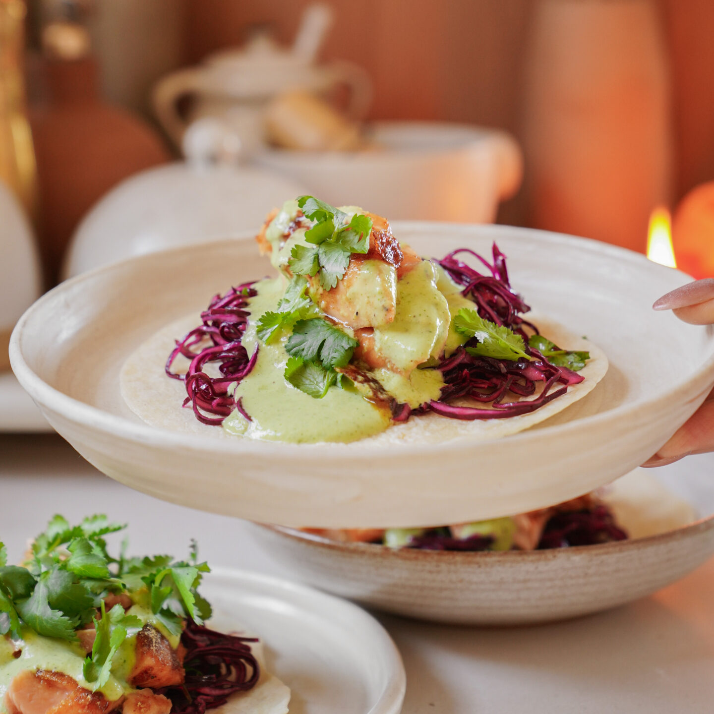 A hand holding a plate with a taco featuring grilled shrimp, purple cabbage, creamy green sauce, and fresh cilantro. The background shows warm-toned kitchenware and a softly lit ambiance.