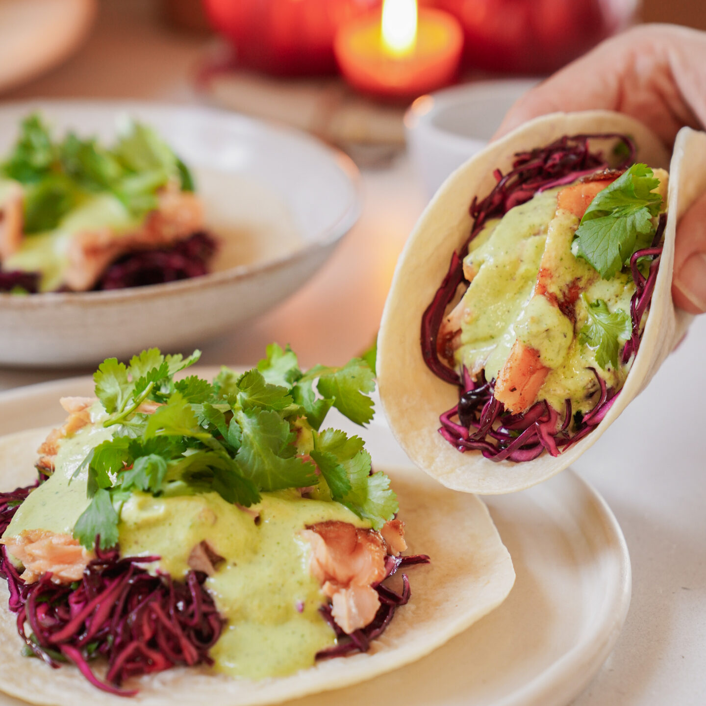 A plate with two soft tacos filled with shredded red cabbage, salmon, green sauce, and garnished with cilantro. A hand holds one taco. A lit candle is blurred in the background, creating a warm ambiance.