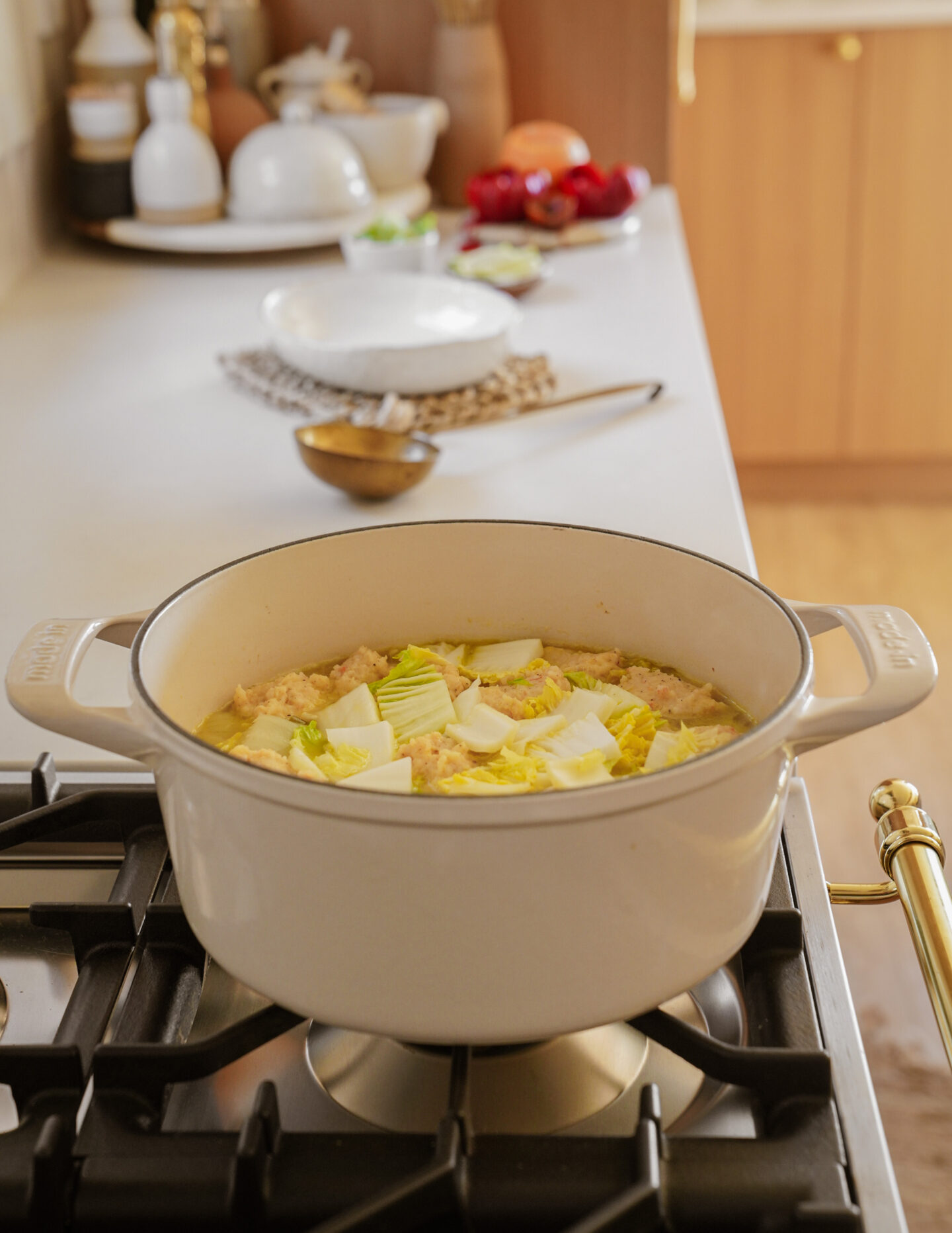 A white pot sits on a stove, filled with a stew containing chunks of cabbage and other ingredients. In the background, a kitchen counter holds various items like bowls, a spoon, and a cutting board with vegetables. The kitchen has warm lighting.