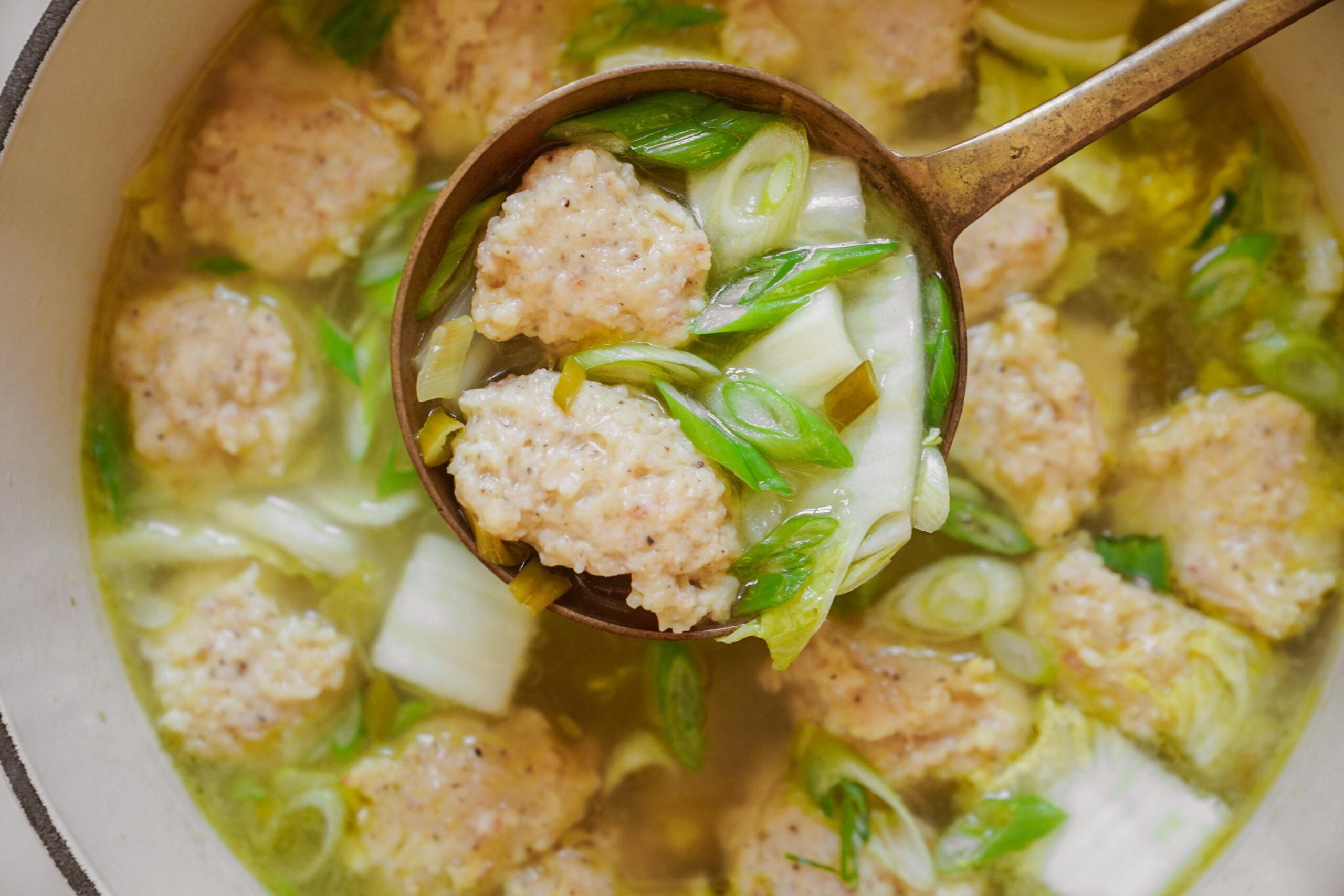 A close-up of a soup featuring meatballs, sliced green onions, and chunks of cabbage. A ladle lifts some of the ingredients from the broth, showcasing the texture of the meatballs and the vibrant green onions.