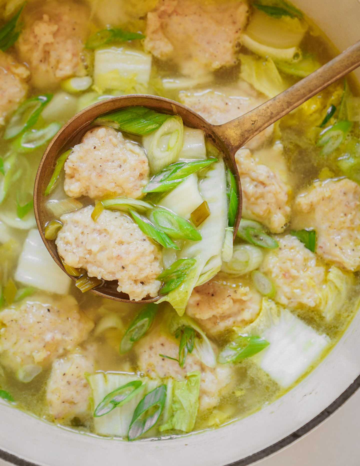 A close-up of a pot containing a clear soup with chicken meatballs, sliced green onions, and cabbage. A ladle lifts some of the ingredients, showcasing the hearty texture of the meatballs and fresh vegetables.