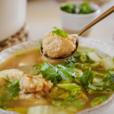 A spoon holding matzo ball above a bowl of matzo ball soup, garnished with fresh cilantro. Background shows a small bowl with more cilantro.