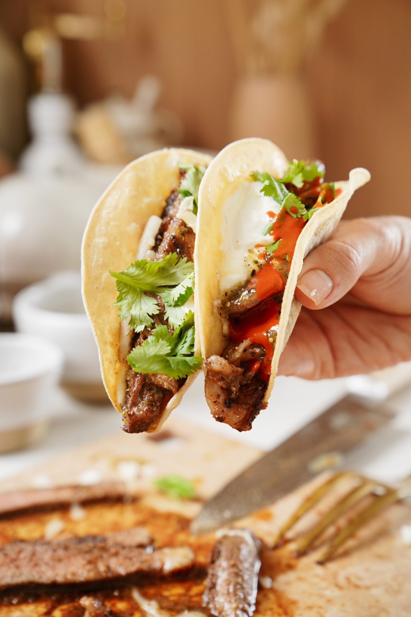 A hand holds two tacos filled with grilled meat, fresh cilantro, sliced red peppers, and a dollop of sour cream. The background features a blurred cutting board with sliced meat and a knife.