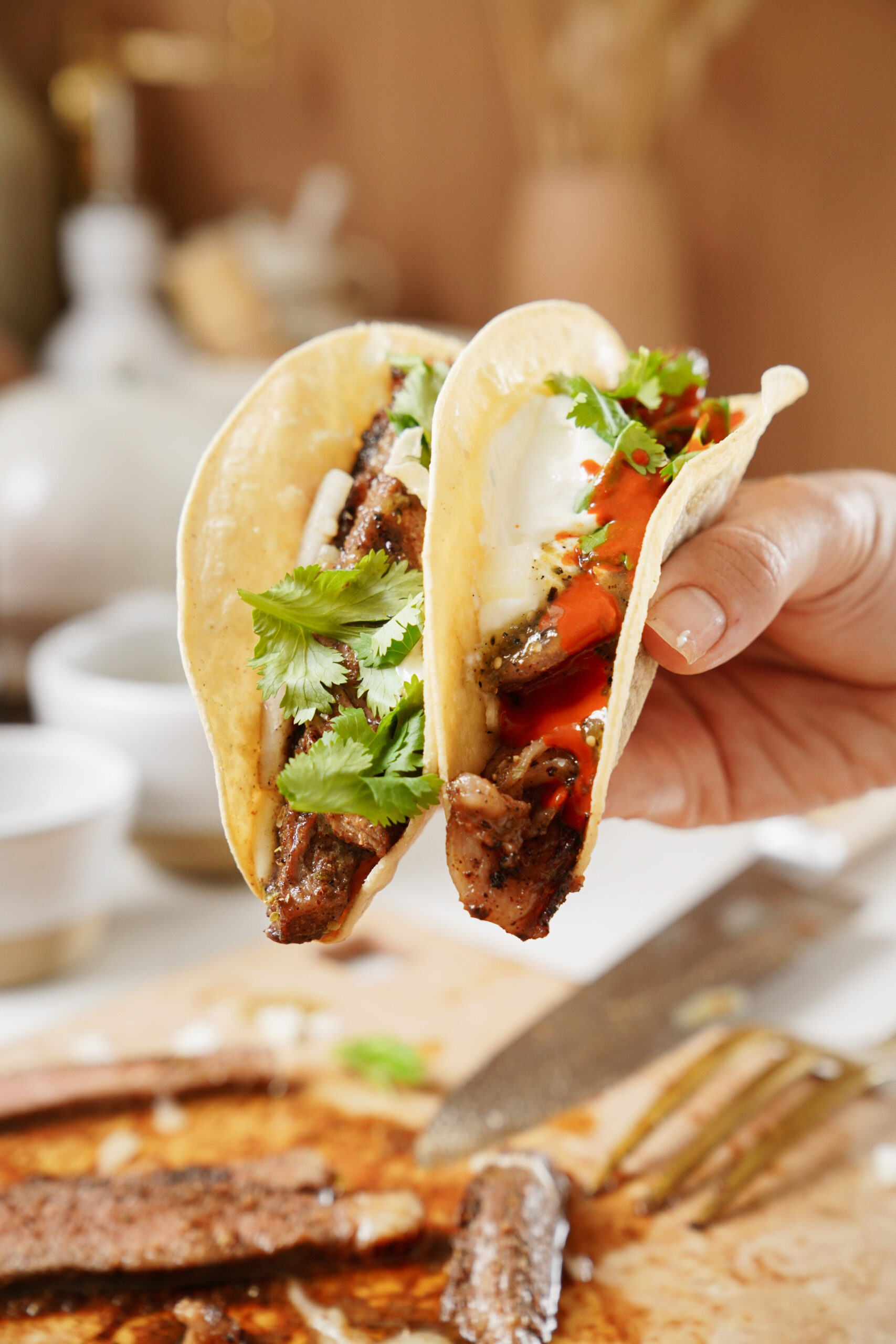 A hand holds two tacos filled with grilled meat, fresh cilantro, sliced red peppers, and a dollop of sour cream. The background features a blurred cutting board with sliced meat and a knife.