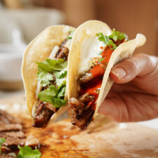 A hand holding two tacos filled with grilled meat, topped with fresh cilantro, onions, and a drizzle of red sauce, wrapped in soft corn tortillas. The background is blurred, focusing on the flavorful tacos.