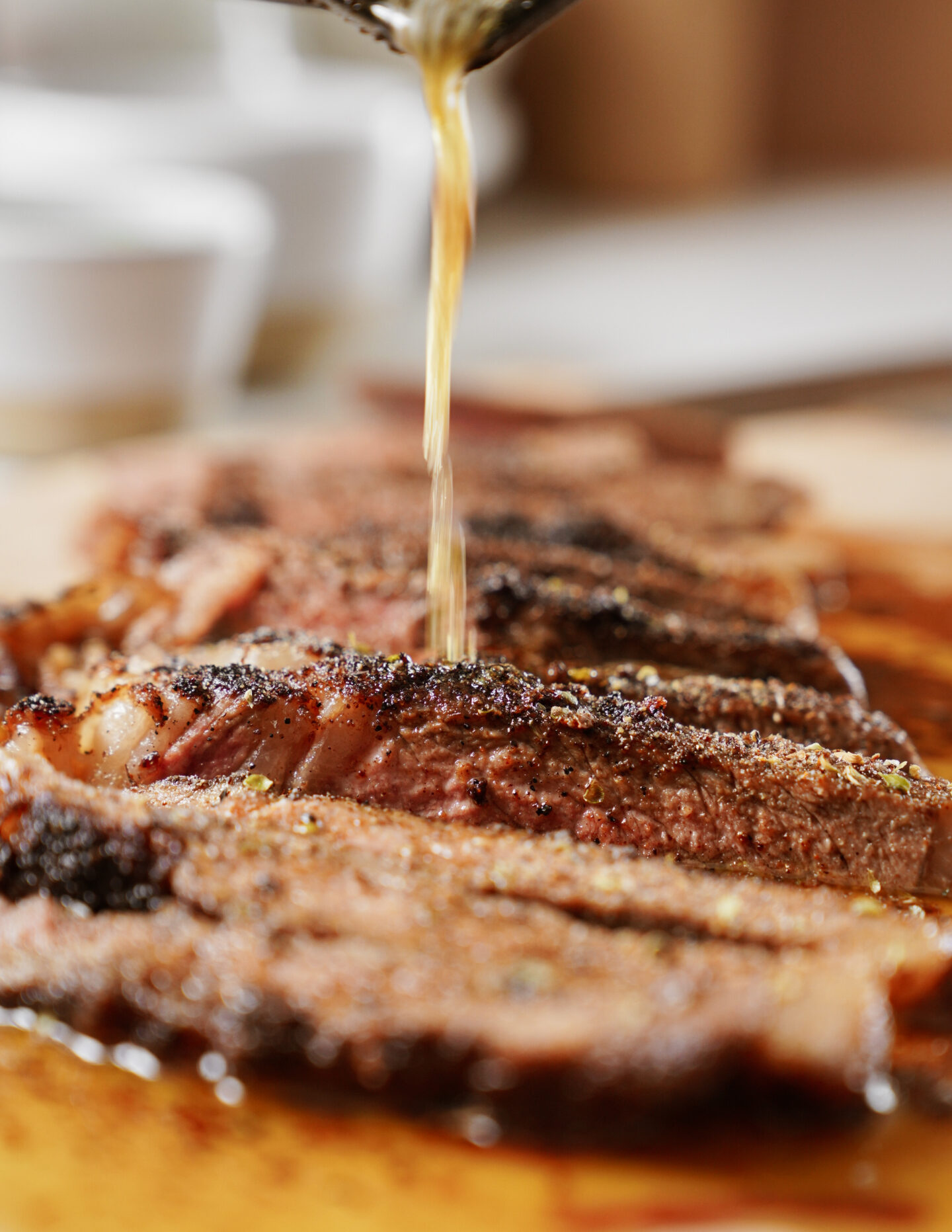 Close-up of sliced, cooked steak on a wooden board, with sauce being poured over the meat. The steak appears juicy and well-seasoned, with a blurred background suggesting a kitchen setting.