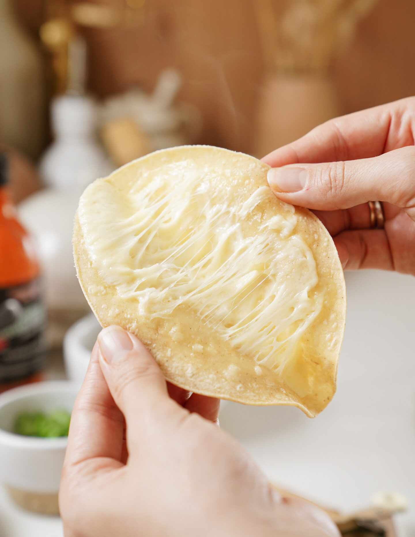 Two hands holding a crispy tortilla with melted cheese pulled apart, showcasing the stretchy texture. The background includes blurred kitchen items, such as a bottle and small bowls, on a white countertop.