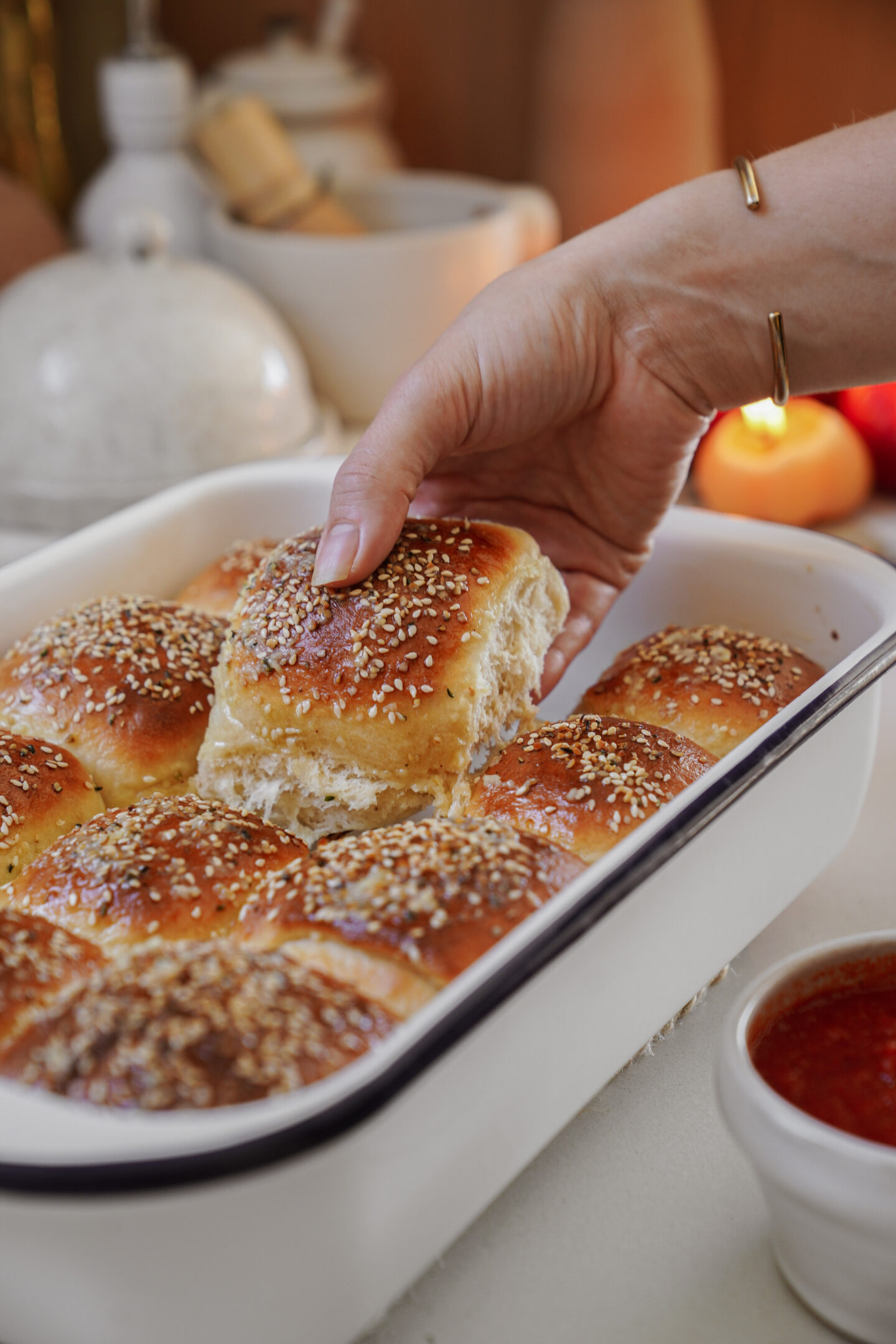 A hand lifts a freshly baked, sesame seed-covered dinner roll from a white baking dish. Several rolls remain in the dish. A small bowl of dipping sauce is nearby, set on a light-colored table.