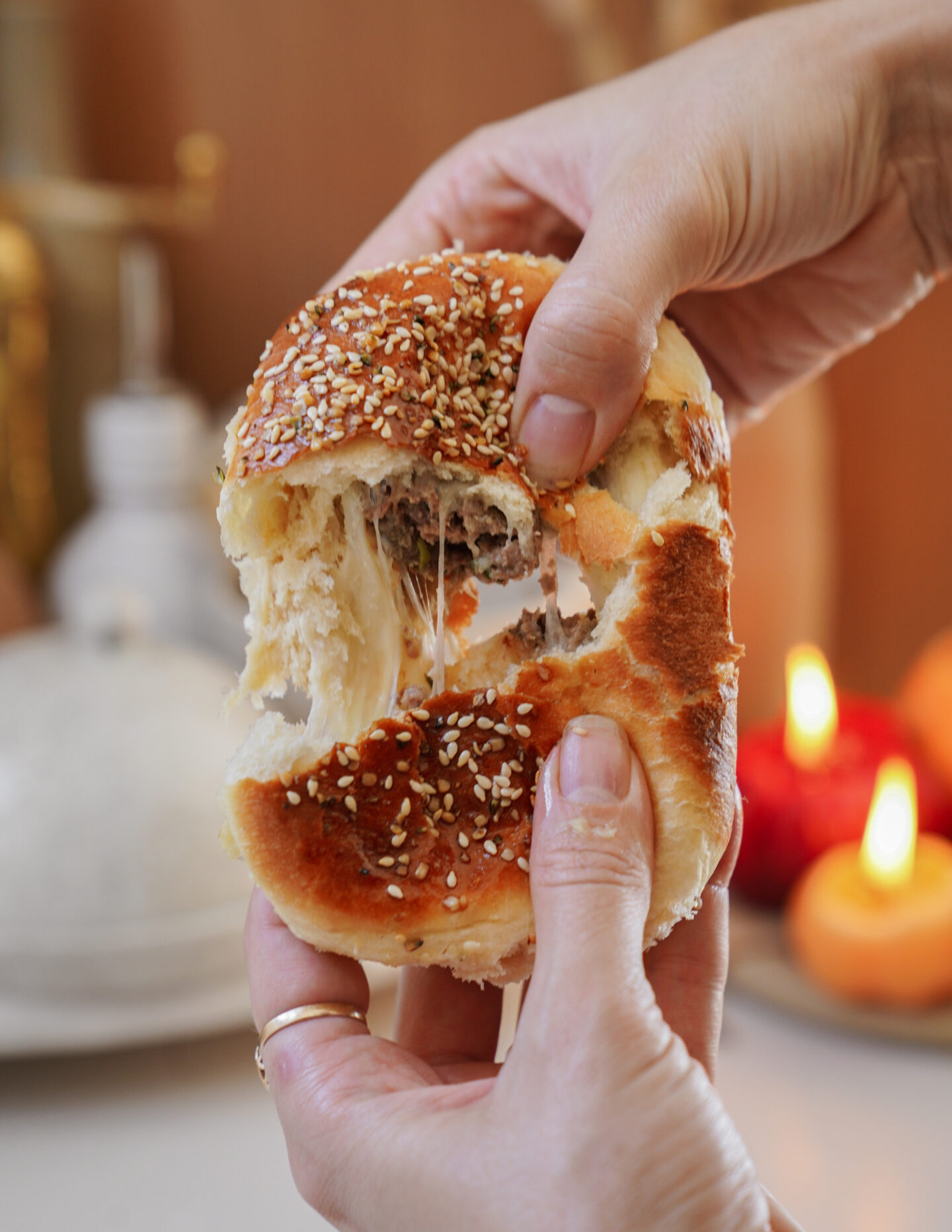 Hands pulling apart a sesame seed-topped bun filled with melted cheese, creating a stringy effect. In the blurred background, there are glowing candles and a white ceramic object.