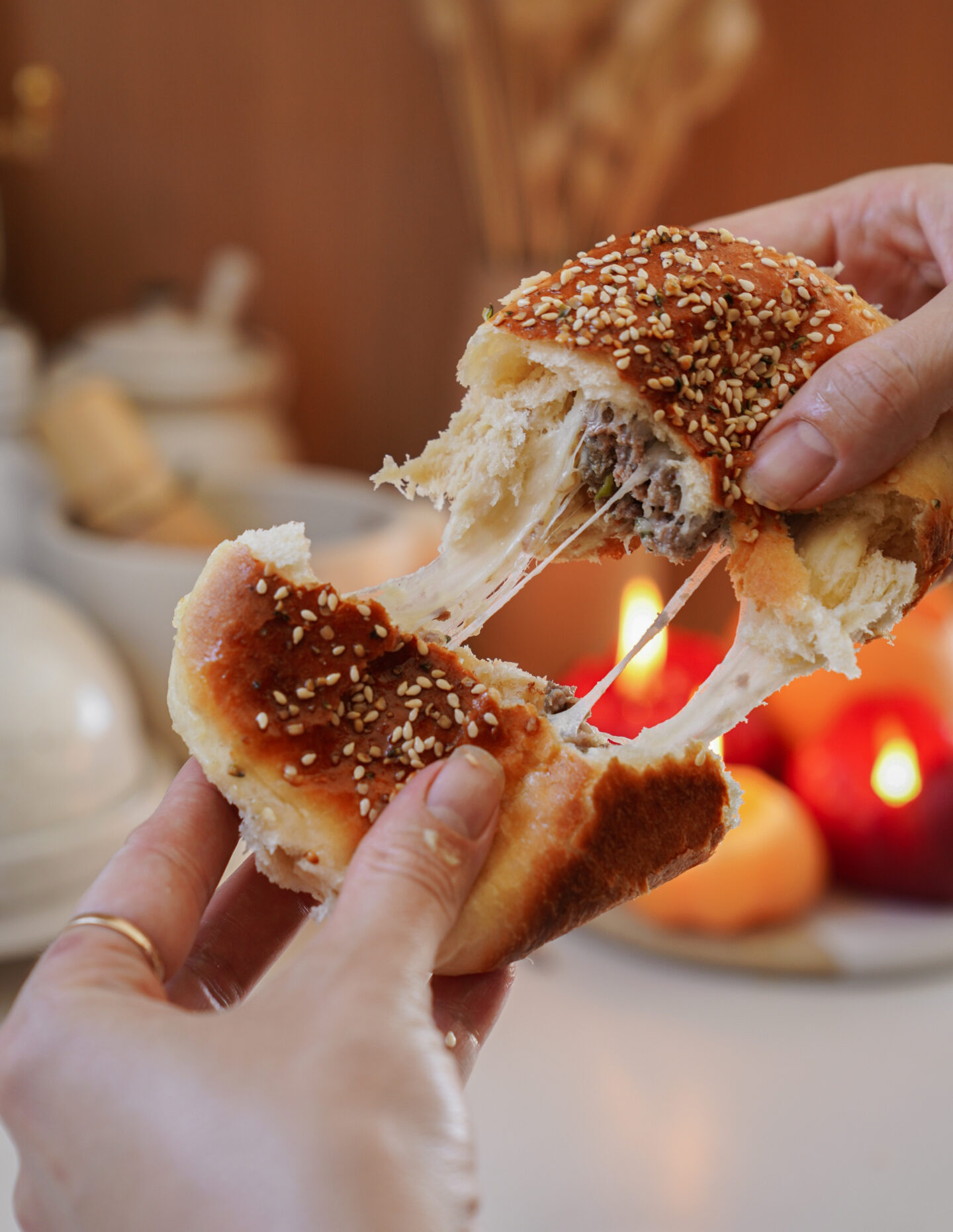 Hands pulling apart a sesame-seed-topped bun, revealing melted cheese inside. A blurred background shows candles and ingredients on a table.